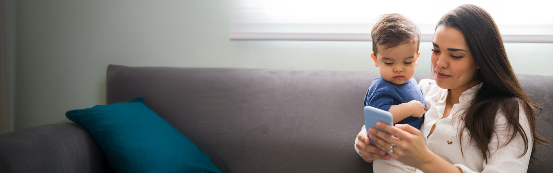 mom with long dark hair sitting on a couch while holding her baby boy and looking at her phone